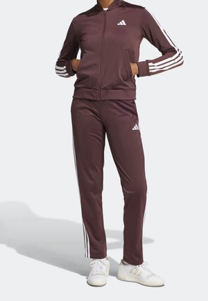 Persoon met een bruin trainingspak met witte strepen op mouwen en broek, staand met handen in jaszakken en witte sneakers.