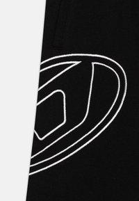 Tissu noir présentant un logo brodé blanc proéminent avec des lignes courbes et des formes géométriques. Une vue rapprochée met en valeur la texture et les détails du design.