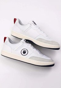 Fehér sneakers, sima bőr felső résszel, szürke velúr részletekkel, kerek logóval, és kontrasztos piros saroktabbal. Gumitalp, mintázott profilú.
