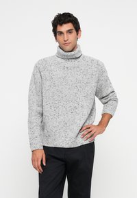 Moški oblečen v sivo speckled pulover s turtleneck ovratnikom in črnimi hlačami, stoji z eno roko na boku ob belem ozadju.