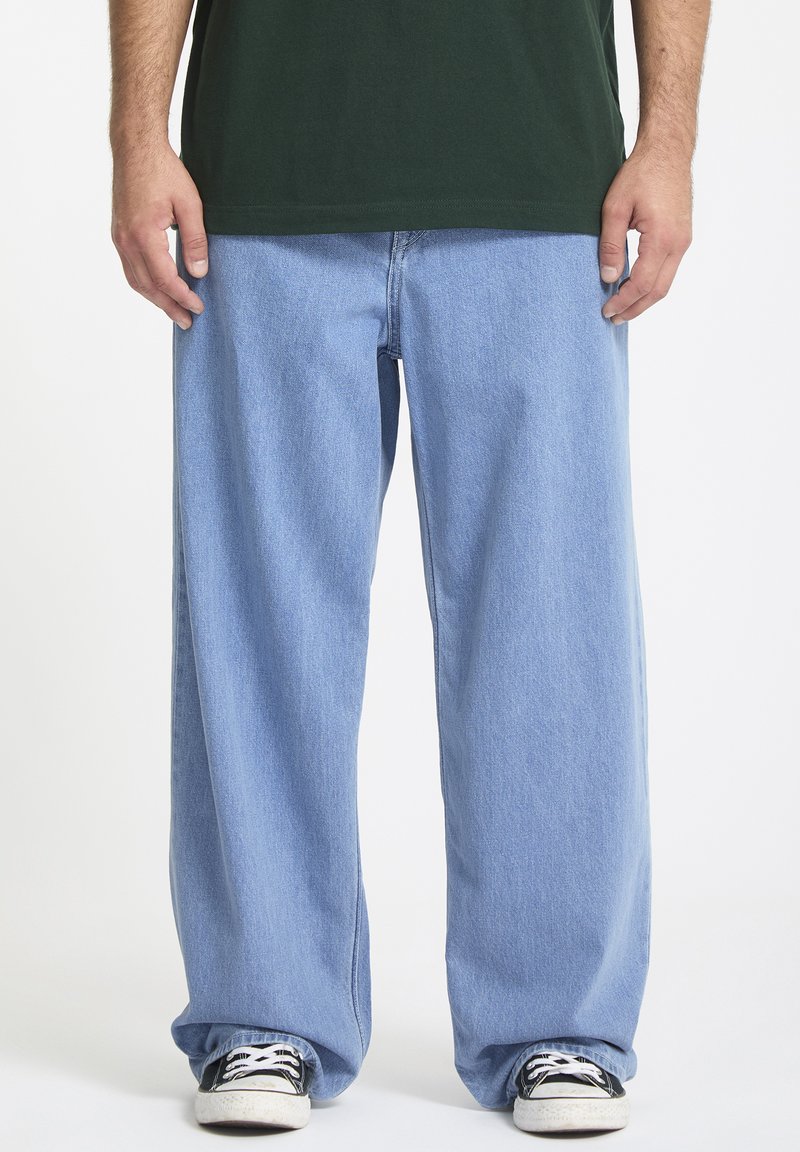 Jean bleu clair à jambes larges en denim, avec une coupe décontractée et associés à des baskets noires. Couleur unie sans motifs visibles.