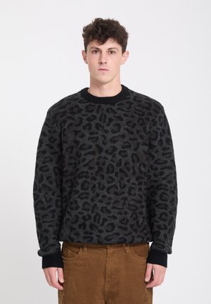 Jeune homme aux cheveux courts et bouclés portant un pull à motif léopard noir et gris foncé et un pantalon en velours côtelé marron, debout sur un fond uni.