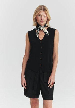 Mujer con cabello rubio lleva una camiseta negra sin mangas con botones, pantalones cortos negros, pañuelo de cuello negro y blanco con estampado y grandes anillos en los dedos.