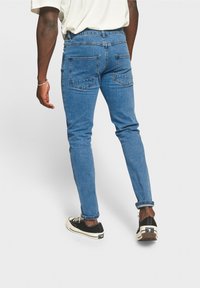 Henkilö, jolla on päällään siniset farkut, valkoinen paita ja mustat matalat lenkkarit, kävelee poispäin yksinkertaista valkoista taustaa vasten, käsi ja rannekoru näkyvissä.