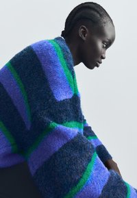 Randig tröja i ullblandning med breda band av lila, blå och grön. Mjuk textur med avslappnad passform, bärs löst på axlarna.