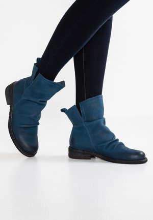 Jambes en jean foncé portant des bottes en cuir bleu décontractées mi-mollet à talons bas sur un sol blanc.