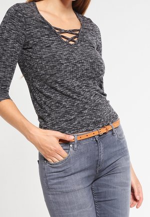 Kvinde iført en mørkegrå krydsdesignet V-halsbluse med trekvartlange ærmer, blå jeans og et tan-farvet bælte med hånden i lommen.