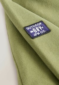 Zelená fleecová látka s jemnou strukturou, obsahující obdélníkovou námořnickou modrou nášivku s nápisem "OUTDOOR DRY JPN CO." v bílém švu.