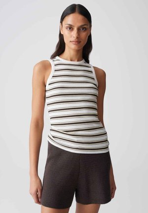 Femme aux longs cheveux foncés portant un haut blanc sans manches avec des rayures horizontales noires et marron ainsi que des shorts marron foncé.