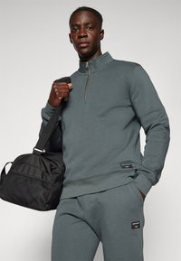 Felpa verde scuro con zip e collo alto, maniche lunghe, dal tocco morbido, abbinata a pantaloni coordinati. Borsa a tracolla nera con dettagli in rete.