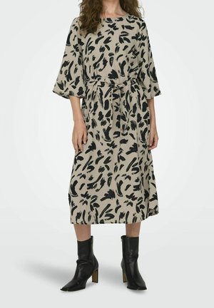 Femme portant une robe midi beige avec des motifs abstraits en coups de pinceau noirs et des bottes en cuir noires à talons mi-mollet, debout devant un fond uni.