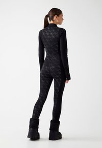 Czarny, całbody strój z teksturowanym wzorem diamentowym na plecach, z długimi rękawami i dopasowanymi nogawkami, w połączeniu z czarnymi, pikowanymi butami.