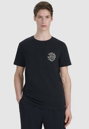 Młody mężczyzna w czarnej koszulce z napisem "Everyday Peace" i okrągłym logo na klatce piersiowej, stoi z jedną ręką w kieszeni.