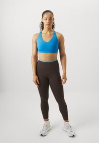 Μπλε αθλητικό σουτιέν με σχέδιο V-λαιμού, σε συνδυασμό με καφέ κολάν ψηλής μέσης. Τα αξεσουάρ περιλαμβάνουν λευκά αθλητικά παπούτσια. Χαρακτηριστικά περιλαμβάνουν απαλό ύφασμα.