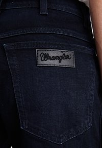 Tamno plave traperice s pravokutnom crnom kožnom etiketom na kojoj piše "Wrangler." Imaju šivene stražnje džepove s oblikom špice.