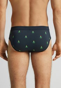 Granatowe slipy kąpielowe z elastycznym pasem, ozdobione drobnymi zielonymi wzorami choinek równomiernie rozmieszczonymi na tkaninie.