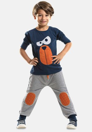 Marineblaues T-Shirt mit einem großen orange-weißen Comic-Hundekopf; graue Sweatpants mit orange karierten Knieflicken und Kordelzug.