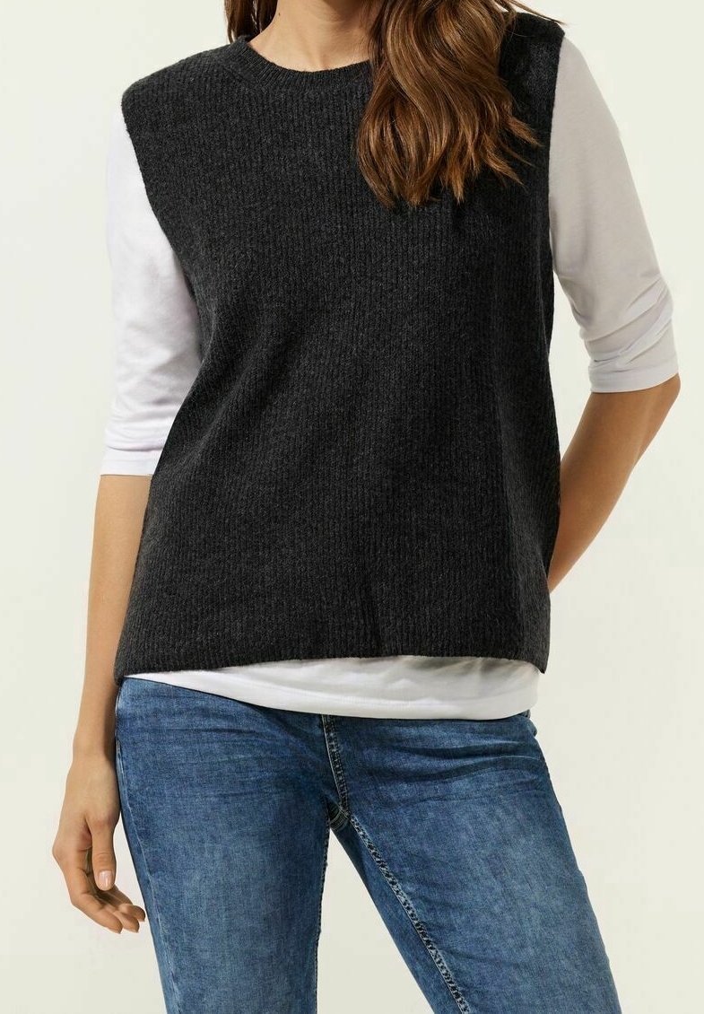 Haut sans manches gris foncé en tricot avec une texture côtelée, porté sur une chemise blanche à manches longues, associé à un jean en denim bleu.
