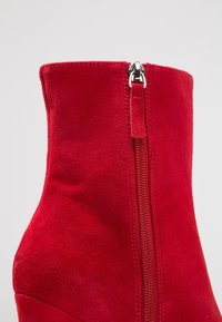 Botín de ante rojo hasta el tobillo, con un caña alta, que presenta una cremallera plateada en el lateral y una textura suave.