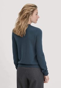 Vrouw met schouderlang blond haar, gekleed in een donkerblauwe trui en grijze krijtstreepbroek, staat zijwaarts tegen een effen achtergrond.