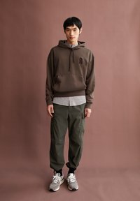 Bruine hoodie met een voorzak en logo, gelaagd over een lichtgrijs shirt, gecombineerd met donkergroene cargobroeken en grijze sneakers.