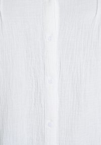 Biela textúrovaná látková košeľa s vertikálnou radou gombíkov a malými okrúhlymi bielymi gombíkmi, detailný pohľad.