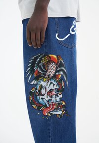 Niebieskie dżinsy z denimu z kolorową, szczegółową haftowaną czaszką i wężem na nogawce, z białym logo na tylniej kieszeni.
