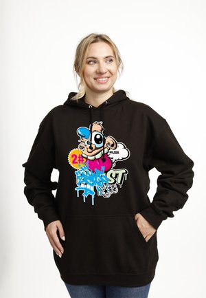 Lächelnde Frau in schwarzem Hoodie mit buntem Cartoon-Gesicht, blauem Graffiti-Schriftzug und einem Abzeichen mit der Nummer "#2", steht mit den Händen in den Taschen.