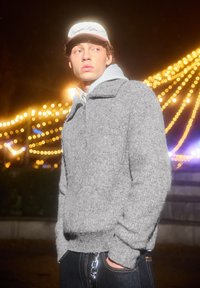 Grauer Strickpullover mit hohem Kragen, kombiniert mit einem hellgrauen Hoodie darunter. Das Modell trägt eine weiße Mütze mit roten Akzenten.