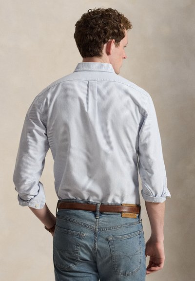 Homme aux cheveux bouclés de dos, portant une chemise à rayures bleu clair boutonnée avec les manches retroussées et un jean bleu avec une ceinture marron.