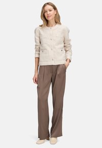 Femme debout, une main dans la poche, portant une veste à boutons crème texturée, un pantalon large marron et des bottines claires.