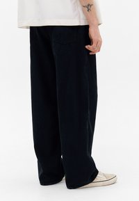 Person iført løse sorte fløjlbukser, blege sneakers og en hvid skjorte, med en fugletattoo synlig på deres underarm.