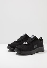 Black athletic shoes made of breathable mesh fabric, featuring round toe, textured sole, and two logo tags on the tongue.
