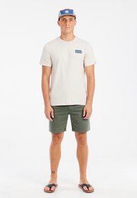 Beige katoenen t-shirt met blauwe grafiek, groene shorts en zwarte flip-flops. Het model draagt een meerkleurige pet met rood, blauw en beige.