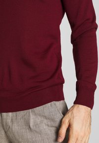 Bordeauxfarbener Pullover mit gerippten Bündchen und Saum, aus einem weichen Strickmaterial gefertigt. Kombiniert mit hellgrauen, strukturierten Hosen.