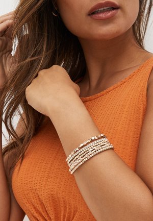 Woman wearing an orange textured sleeveless top shows multiple gold and beaded bracelets on her wrist, touching her hair with the other hand.
