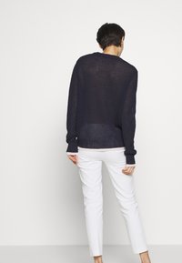 Person mit kurz dunklem Haar, der einen lockeren schwarzen Strickpullover und weiße Hosen trägt, steht vor einem schlichten hellen Hintergrund und ist abgewandt.