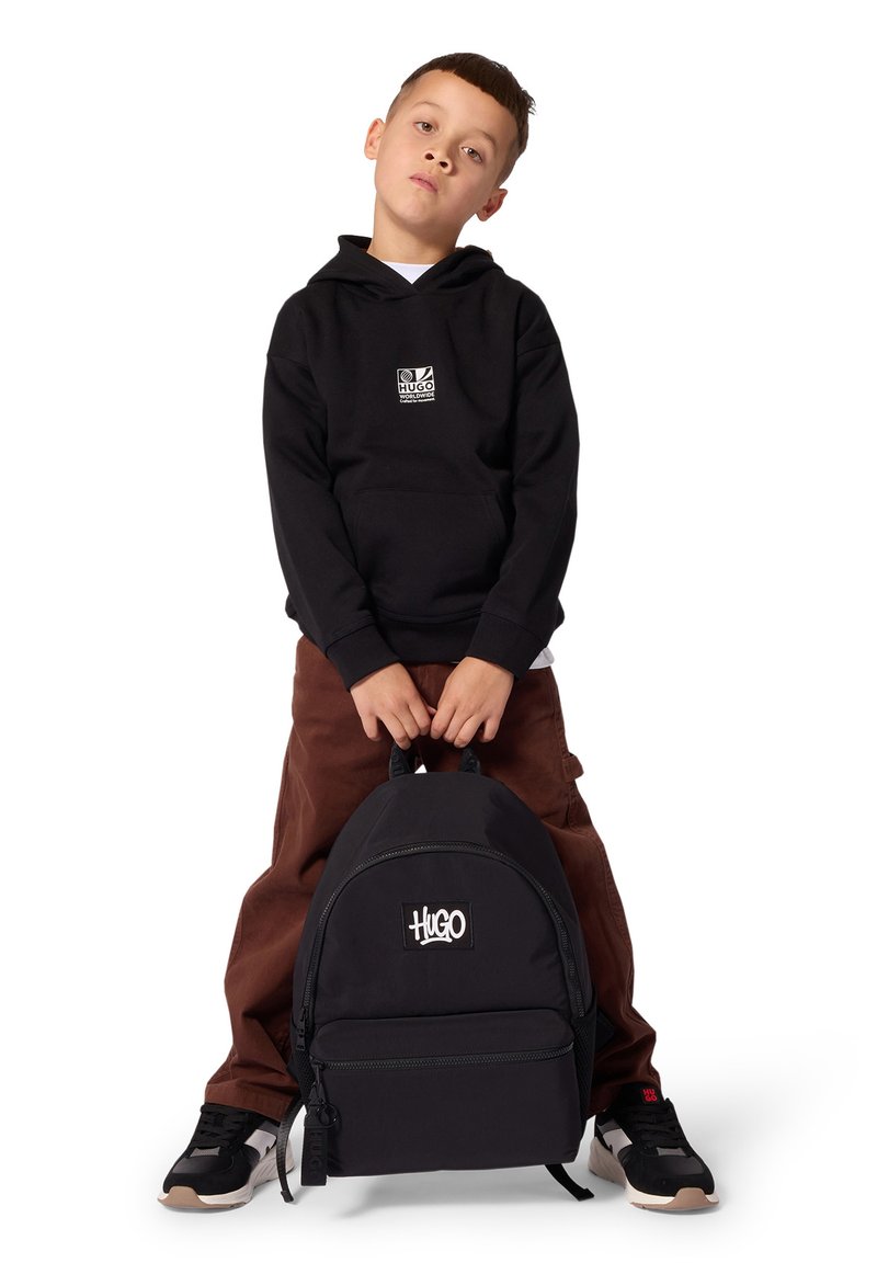Νέος αγόρι φοράει μαύρο hoodie και καφέ παντελόνια κρατάει μπροστά του μαύρο σακίδιο πλάτης Hugo ενώ στέκεται μπροστά σε λευκό φόντο.