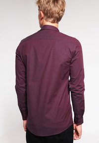 Chemise bordeaux à manches longues avec un motif géométrique subtil, col boutonné et texture lisse. Coutures et poignets visibles.