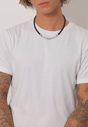 Sort flettet halskæde med grønne perler og sølvdetaljer, båret over en enkel hvid t-shirt, der fremhæver et afslappet, lagdelt look.