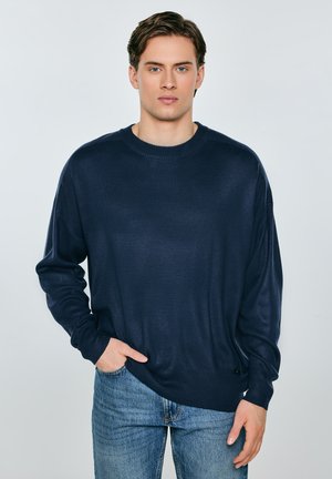 Młody mężczyzna o jasnej karnacji i brązowych włosach, ubrany w granatowy sweter i niebieskie dżinsy, stojący z jedną ręką w kieszeni na jednolitym tle.