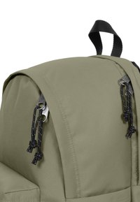 Zelený látkový batoh s zaobleným tvarem, černými akcentovými popruhy, kovovými komponenty a stahováním na šňůrku. Texturovaný, odolný materiál.