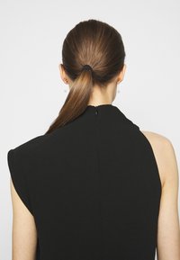 Svart ärmlös plagg med hög halsringning och dragkedjedetalj bak, med slät, lätt strukturerad tyg och subtil axelstruktur.