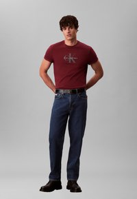 Burgundia värvi T-särk Calvin Kleini logoga, kombineeritud siniste teksapükstega ja mustade saapadega. Modell seisab käed puusal, neutraalse taustaga.