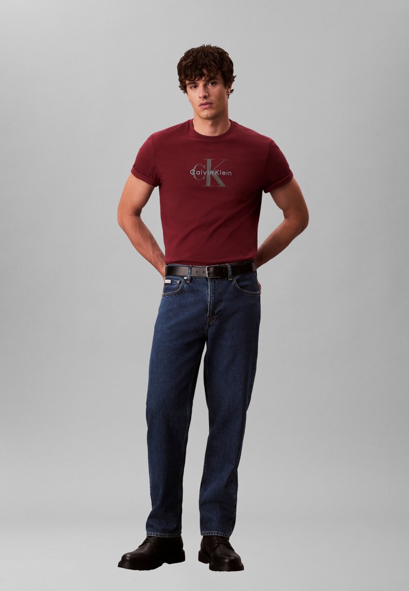 Burgundia värvi T-särk Calvin Kleini logoga, kombineeritud siniste teksapükstega ja mustade saapadega. Modell seisab käed puusal, neutraalse taustaga.