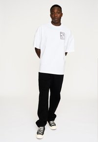 Hvid oversized t-shirt med et grafisk tryk på venstre bryst, kombineret med sorte bukser og sort-hvide sneakers. Enkel og ren design.