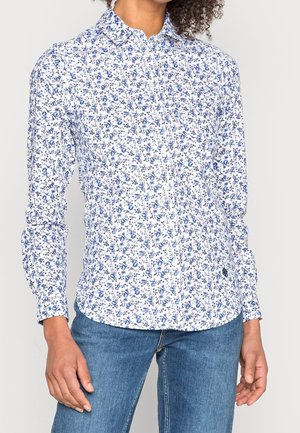 Mujer que lleva una camisa blanca de manga larga con botones, con un estampado floral azul y vaqueros de mezclilla azules.