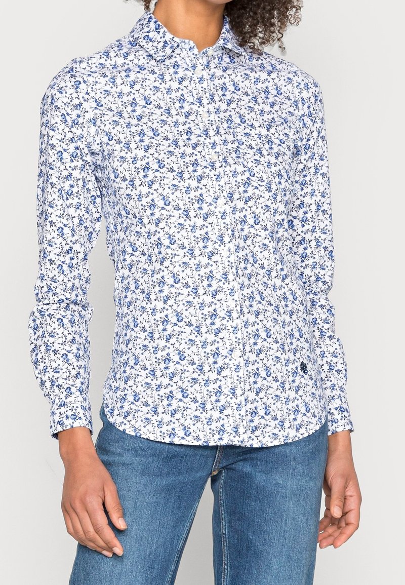 Femme portant une chemise blanche à manches longues boutonnée avec un motif floral bleu et un jean en denim bleu.