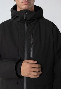 Svart huva jacka med dragkedja fram och ficka med dragkedja på bröstet. Har en slät textur, elastiska ärmslut och logotypdetalj på bröstet.