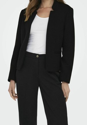 Vrouw draagt een zwarte gestructureerde blazer over een witte top, gecombineerd met een zwarte broek met hoge taille tegen een effen lichte achtergrond.